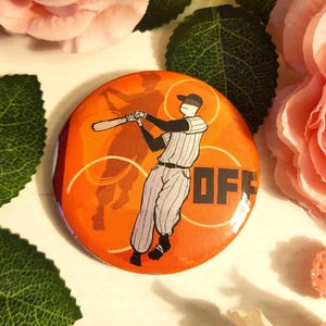 May include: Circular orange button featuring a baseball player illustration. The player is swinging a bat, with a shadow of the same image behind it. The word "OFF" is printed in black. The button is surrounded by pink flowers and green leaves.