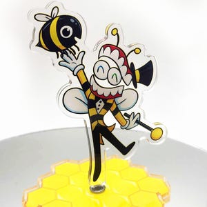 Puede incluir: Un adorno para pastel de acrílico caprichoso con un personaje de abeja de dibujos animados con un traje a rayas negro y amarillo, sosteniendo una abeja de dibujos animados. El adorno está colocado sobre una base de panal amarilla. El personaje de la abeja lleva un sombrero rojo y blanco y un sombrero de copa negro.