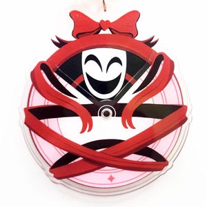 May include: A round, clear acrylic charm featuring a smiling mask design. The mask is white with a black outline and a wide grin. Red ribbons and a bow with a heart accent the design. The charm has a pink and white background with a diamond shape.