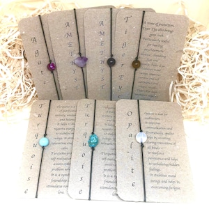 Pode incluir: Um conjunto de oito pulseiras com pedras e contas de várias cores. Cada pulseira tem um cartão anexado com o nome da pedra e suas propriedades. As pedras incluem turquesa, ametista, olho de tigre e opala.