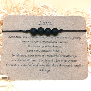 Puede incluir: Pulsera de piedra de lava negra con un cordón negro. La pulsera tiene cinco cuentas y el texto "Lava" está impreso en una tarjeta marrón. La tarjeta también incluye información sobre los beneficios de la piedra de lava.