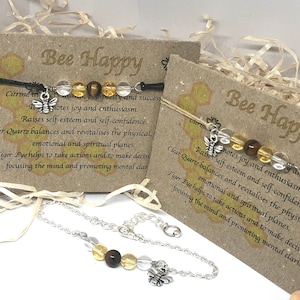 May include: Two bracelets with silver chains and bee charms. The bracelets feature clear quartz, tiger eye, and citrine beads. The cards next to the bracelets have the text "Bee Happy" and descriptions of the stones and their properties.