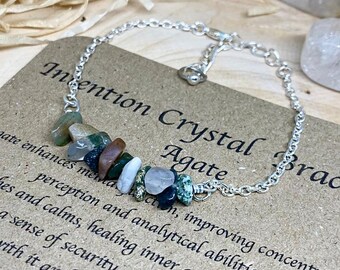 May include: A silver chain bracelet with a charm made of multiple small, colorful agate stones. The bracelet is on a brown card with the text "Intention Crystal Bracelet Agate" and a description of the stone's properties.