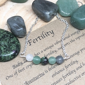 May include: A silver chain bracelet with seven beads, including a white, a green, a pink, a green, a grey, and two green beads. The bracelet is on a brown surface with the text "Fertility" and "Ruby Zo" written on it.