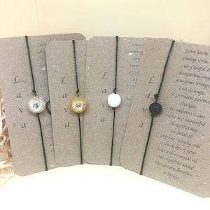 May include: Four black string bracelets with lava stone beads. The beads are black, white, and gold. Each bracelet has a card attached with text about the benefits of lava stones. The text on the card reads "Lava Stone calming stone, very useful in dissipating anger and gives strength and courage. It promotes positive changes. In addition, Lava Stone is a wonderful aromatherapy essential oil diffuser. Simply add a few drops of your favourite essential oil and enjoy the added therapeutic benefits it brings."