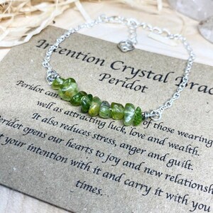 Op de afbeelding: Een zilveren armband met een rij kleine, levendige groene peridotstenen. De armband wordt getoond op een kaart met de tekst "Intention Crystal Bracelet Peridot". De kaart bevat ook tekst over de eigenschappen van de peridot.
