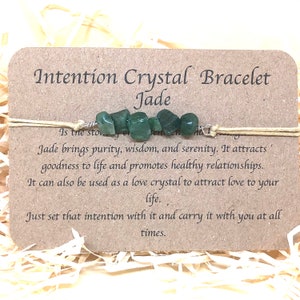 Op de afbeelding: Een groene jadekristallen armband met een beige koord. De kaart vermeldt "Intention Crystal Bracelet Jade" en bevat een beschrijving van de eigenschappen van de steen.