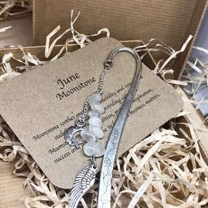 Puede incluir: Un marcapáginas de plata con un amuleto de piedra lunar de junio. El marcapáginas presenta un ala de plata y un diseño floral. El amuleto tiene una luna, una estrella y una luna creciente. El texto en la tarjeta dice "Junio Piedra lunar. La piedra lunar calma las emociones, la estabilidad y el estrés, proporcionando calma, intuición, prosperidad, inspiración y éxito y asuntos comerciales positivos."