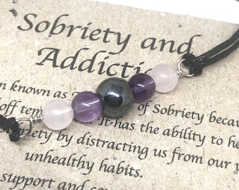 May include: A black cord bracelet with amethyst, rose quartz, and hematite beads. The bracelet is on a card with the text "Sobriety and Addiction".