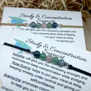 Può includere: Un braccialetto in corda nera con quattro perle di pietre preziose: sodalite, quarzo rosa, aventurina e sodalite. Il braccialetto è accompagnato da una carta che recita "Study & Concentration Support" e elenca le proprietà di ogni pietra preziosa. La carta include anche il testo "ArtemisIdeas".
