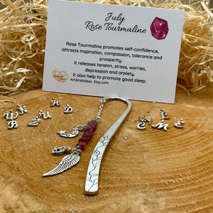 May include: Silver bookmark featuring a rose tourmaline charm, a crescent moon and star charm, an angel wing charm, and a chain with small beads. The bookmark is on a wooden surface with a card that reads "July Rose Tourmaline" and describes the properties of the stone.