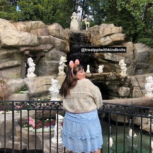 Puede incluir: Una mujer con un su&eacute;ter de crochet colorido y una falda azul claro se encuentra frente a una cascada de piedra con siete peque&ntilde;as estatuas blancas de enanos. El texto "@treatpplwithkindknits" es visible en la roca.