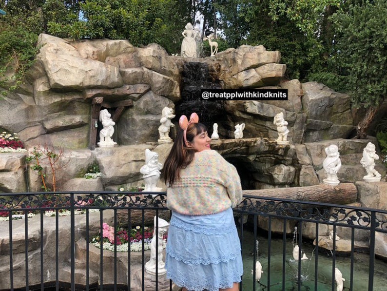 Puede incluir: Una mujer con un su&eacute;ter de crochet colorido y una falda azul claro se encuentra frente a una cascada de piedra con siete peque&ntilde;as estatuas blancas de enanos. El texto "@treatpplwithkindknits" es visible en la roca.