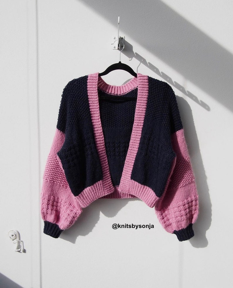 Puede incluir: Un c&aacute;rdigan de punto rosa y azul marino con cuello en V y mangas largas. El c&aacute;rdigan est&aacute; colgado de una percha contra una pared blanca. El texto "@knitsbysonja" es visible en la esquina inferior derecha de la imagen.
