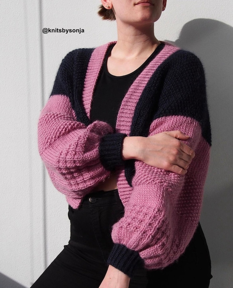 Puede incluir: Un c&aacute;rdigan de punto rosa y azul marino con cuello en V y mangas largas. El c&aacute;rdigan tiene un patr&oacute;n de punto de cable en las mangas y un patr&oacute;n de punto liso en el cuerpo. El c&aacute;rdigan es corto y tiene un ajuste holgado. @knitsbysonja
