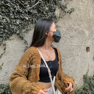 Puede incluir: Una mujer lleva un c&aacute;rdigan de punto marr&oacute;n con un top negro de tirantes y pantalones blancos. Lleva una mascarilla verde oscuro y un collar de plata con un colgante de coraz&oacute;n. El texto "@iam_janka" es visible en el c&aacute;rdigan.