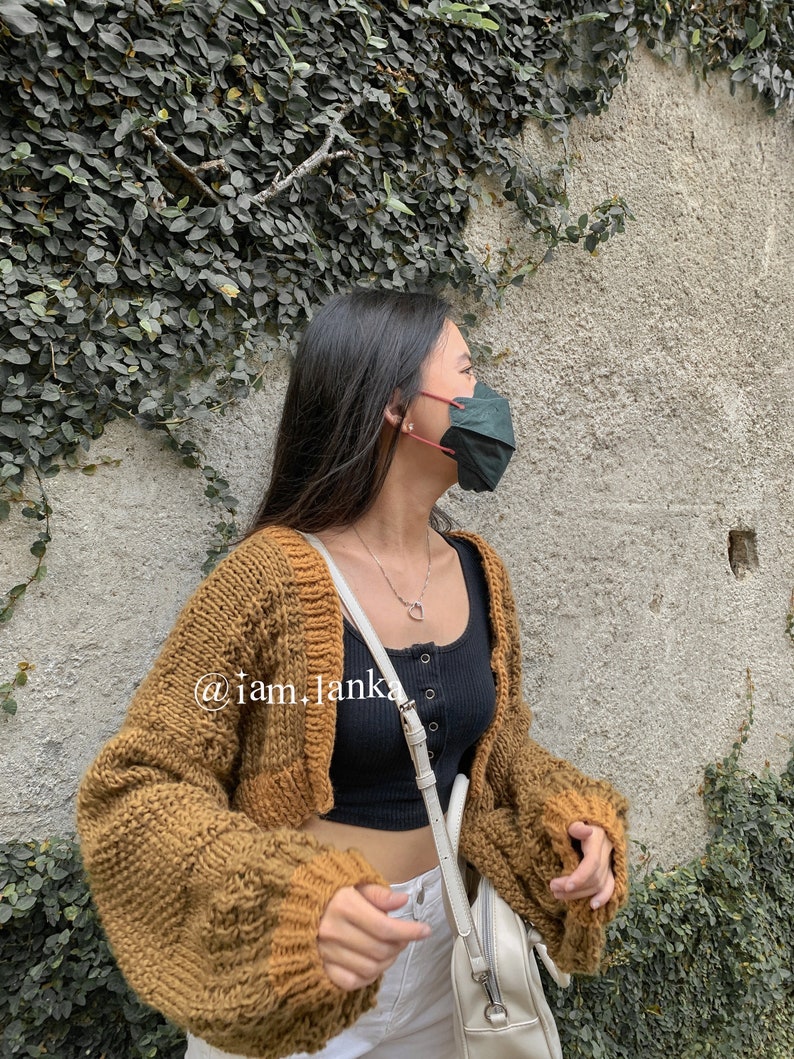 Puede incluir: Una mujer lleva un c&aacute;rdigan de punto marr&oacute;n con un top negro de tirantes y pantalones blancos. Lleva una mascarilla verde oscuro y un collar de plata con un colgante de coraz&oacute;n. El texto "@iam_janka" es visible en el c&aacute;rdigan.
