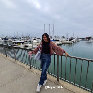 Puede incluir: Una persona con un c&aacute;rdigan multicolor, una camisa negra y pantalones vaqueros azules, de pie en un muelle con una barandilla y vista a un puerto con barcos. La leyenda en la parte inferior de la imagen dice "@theyarnbitch".