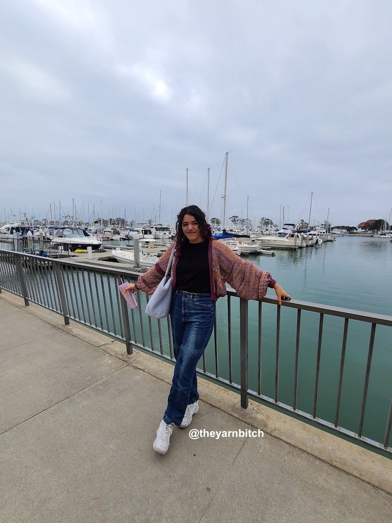 Puede incluir: Una persona con un c&aacute;rdigan multicolor, una camisa negra y pantalones vaqueros azules, de pie en un muelle con una barandilla y vista a un puerto con barcos. La leyenda en la parte inferior de la imagen dice "@theyarnbitch".