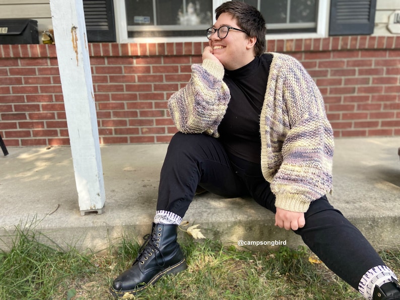Puede incluir: Una persona lleva una camisa negra de cuello alto, pantalones negros y un c&aacute;rdigan multicolor. Lleva botas negras con calcetines blancos y gafas. La persona est&aacute; sentada en un porche con una pared de ladrillo en el fondo. El texto "@campsongbird" es visible en la imagen.