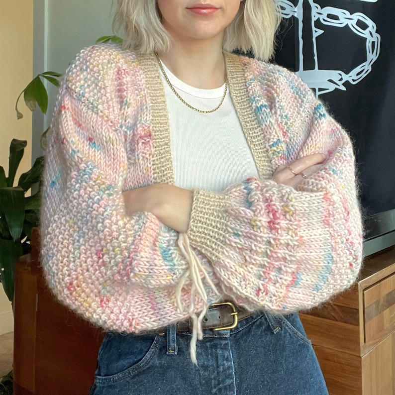 Puede incluir: Un c&aacute;rdigan de punto rosa, azul y amarillo con un lazo en la mu&ntilde;eca. El c&aacute;rdigan tiene un corte holgado y est&aacute; hecho de un hilo suave y peludo.