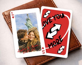 May include: Two custom playing cards with a brown leather wallet. One card features a couple kissing in front of the Statue of Liberty. The other card has the words "LOVE YOU MORE" with arrows on a red background.