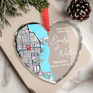 May include: Heart-shaped glass Christmas ornament with a map design and the text "OUR FIRST CHRISTMAS Emily Nathan ENGAGED October 12th 2025". The ornament has a red ribbon for hanging.