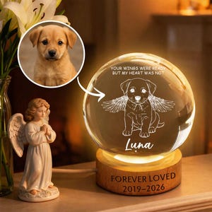 Könnte beinhalten: Eine Gedenk-Kristallkugel mit einer Hundeillustration und dem Text "Your wings were ready but my heart was not". Auf der Basis steht "Forever Loved 2019-2026". Ein Foto eines Welpen befindet sich in einem Kreis. Eine Engelsstatue ist im Vordergrund.