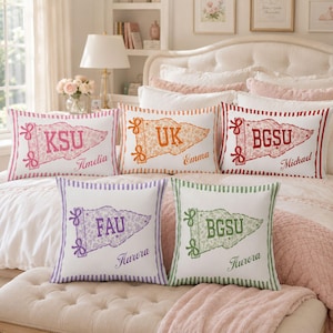 May include: Five white rectangular throw pillows with striped edges, each featuring a pennant graphic with a university acronym and a personalised name. The pennants are printed in pink, orange, red, purple, and green with floral patterns.