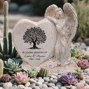 May include: A heart-shaped memorial stone with an angel statue. The stone features a black tree design and the inscription "IN LOVING MEMORY OF Laura R. Hunter 1960-2026." The angel is sculpted with detailed wings. The scene is set among succulents and pebbles.