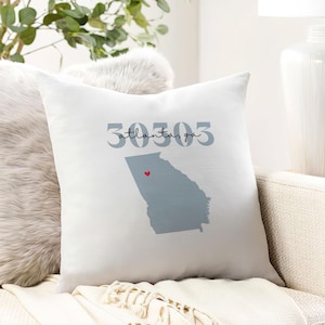 May include: White square throw pillow featuring a gray outline of the state of Georgia with a red heart marking a location. The text "30303 Atlanta, ga" is printed above the state outline. The pillow is on a beige sofa.