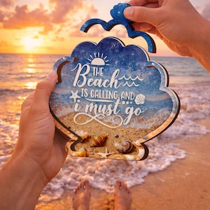 May include: A wooden beach-themed sign with the text "The beach is calling, and I must go." The sign features a blue sky and ocean design with seashells and a starfish. A blue decorative piece is held above the sign.