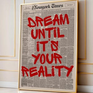 以下が含まれることがあります： ヴィンテージ新聞を背景に、「DREAM UNTIL IT'S YOUR REALITY」という赤い文字が書かれた額入りのプリント。プリントは金のフレームに入っています。モチベーションを高めるアート作品。