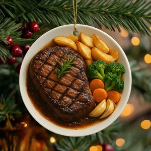 May include: A round, white ceramic ornament featuring a grilled steak with a rosemary sprig, surrounded by roasted potatoes, broccoli, carrots, and garlic cloves. The ornament is suspended by a gold string against a backdrop of Christmas greenery.