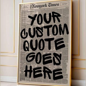 以下が含まれることがあります： 新聞の背景に「YOUR CUSTOM QUOTE GOES HERE」というテキストが黒い太字で書かれた額入りのプリント。フレームは金色で、プリントはニュートラルな壁に飾られています。