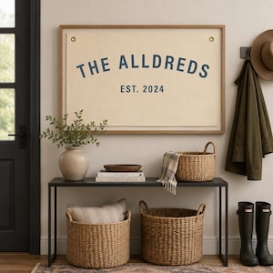 May include: A rectangular wall hanging featuring a cream-colored fabric banner with brass grommets in a wooden frame. The text "THE ALLDREDS" is printed in a dark blue arched font above the text "EST. 2024" in a matching dark blue color.