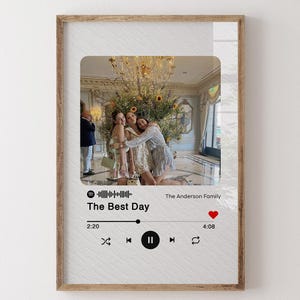May include: A framed print with a photograph of three people hugging, set in a lavish interior. The design includes a Spotify-style interface with the song title "The Best Day" and a heart icon, all within a wooden frame.