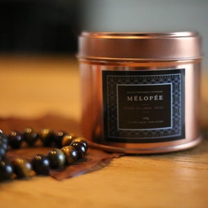 Peut inclure: Un pot de bougie en cuivre avec une étiquette noire sur laquelle est inscrit "MÉLOPEE" et "BOIS DE CÈDRE, MUSC". La bougie est posée sur une surface en bois avec un bracelet en perles.