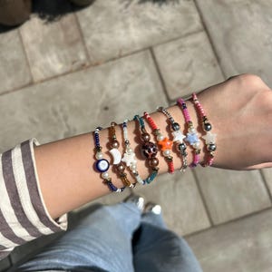 May include: A selection of beaded bracelets in diverse colours and designs. The bracelets incorporate white, blue, pink, and gold beads, along with star, moon, and eye charms. The bracelets are displayed on a wrist, with a striped shirt and jeans in the background.