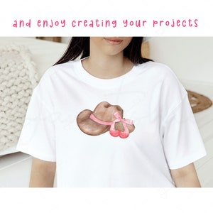 以下が含まれることがあります： 白いTシャツに、茶色のカウボーイハットとピンクのリボン、そしてその上に赤いチェリーが2つ乗っています。