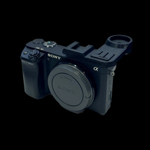 Può includere: Una fotocamera Sony Alpha nera con un copriobiettivo e un accessorio nero attaccato alla parte superiore.