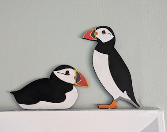 May include: Two flat, cutout-style puffin figures. One puffin is standing upright with orange feet, while the other is in a resting, prone position. Both feature black and white plumage with distinctive colorful beaks in red, yellow, and black.