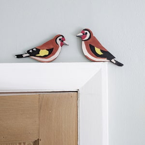 Peut inclure: Deux décorations d'oiseaux en bois. Chaque oiseau a un corps brun, une aile blanche et noire, et une tête rouge, blanche et noire. Les oiseaux sont posés sur un cadre de porte blanc contre un mur bleu clair.