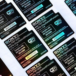 May include: Close-up of multiple holographic stickers for the Sony PlayStation Portable (PSP) handheld gaming console. The stickers are black with white text and a Wi-Fi certified logo. The text on the stickers reads "Model PSP-2000 PSP (PlayStation®Portable) DC 5V 1.2A Complies with IDA Standards DB102988" and "SERIAL No. HE2127333 MADE IN CHINA".