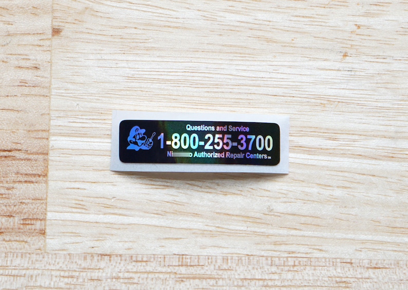 May include: A holographic sticker featuring a cartoon character holding a spanner. The sticker reads "Questions and Service 1-800-255-3700 Nintendo Authorised Repair Centres."