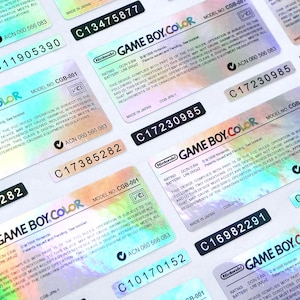 Puede incluir: Una colección de etiquetas holográficas de Game Boy Color con números de serie. Cada etiqueta presenta el logotipo de Nintendo y el texto "GAME BOY COLOR". Las etiquetas tienen un brillo de arcoíris y son rectangulares.