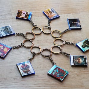 May include: A collection of miniature video game case keychains. Each keychain features a game cover design, attached to a chain and a gold-colored ring. The keychains are arranged in a circular pattern on a wooden surface.