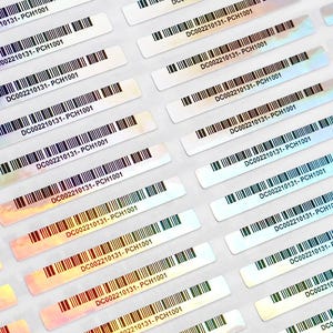 以下が含まれることがあります： 複数のホログラフィックバーコードステッカーのクローズアップ。各ステッカーには、固有のバーコードとテキスト「DC002210131-PCH1001」があります。ステッカーは、ピンク、青、黄色などの色を反射する、光沢のある虹色の仕上がりになっています。