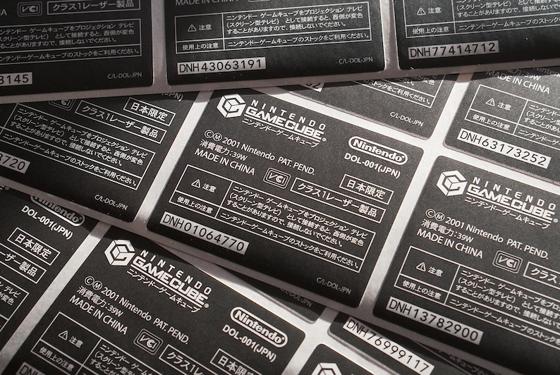 May include: Close-up of multiple black and white Nintendo GameCube labels with the text "NINTENDO GAMECUBE" and "MADE IN CHINA". The labels also include the text "DOL-001(JPN)" and "CLASS 1 LASER PRODUCT".