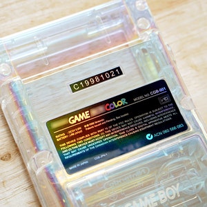 May include: A clear Game Boy Color handheld gaming console with a gold label that reads "GAME COLOR" and includes the model number "CGB-001". The label also includes the rating, battery, and other information.